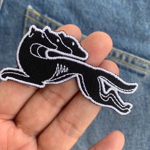 Puede incluir: Parche bordado en blanco y negro con una representación estilizada de tres perros en posición de carrera. El parche está cosido sobre un fondo de mezclilla, mostrando los colores contrastantes y las costuras detalladas.