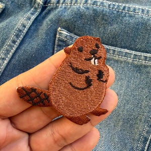 Może przedstawiać: Brązowa, haftowana naszywka z bobrem z czarnymi detalami, w tym ogonem, łapami i rysami twarzy. Naszywka jest trzymana na tle dżinsu.