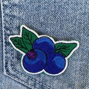 以下が含まれることがあります： 青いデニム生地に、緑の葉が付いたブルーベリー3つが刺繍されたパッチが縫い付けられている。