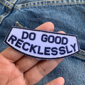 Puede incluir: Un parche morado claro con las palabras "DO GOOD RECKLESSLY" bordadas en azul marino. El parche tiene un borde blanco y está sobre un fondo de vaqueros azules.