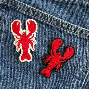 May include: Two embroidered lobster patches featuring bright red thread. One patch has a cream-coloured border, while the other has a black border. Both patches display detailed stitching of the lobster's claws, legs, and segmented tail, creating a textured appearance.