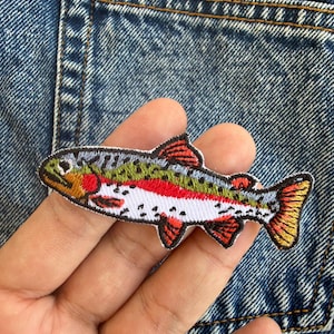 Puede incluir: Parche bordado con un diseño de pez trucha colorido. El parche tiene un borde blanco y muestra una representación detallada del pez con acentos rojos, verdes y amarillos. Ideal para añadir a chaquetas, bolsos u otros artículos de tela.