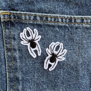 May include: Two embroidered spider patches featuring black bodies and legs outlined in white thread. The patches are positioned on the dark blue denim fabric of a pocket, showcasing detailed stitching and a high-contrast design on the textured material.