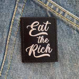 Op de afbeelding: Zwarte geborduurde patch met witte tekst die "Eat the Rich" zegt op een denim achtergrond.