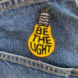 Puede incluir: Un parche amarillo en forma de bombilla con el texto "BE THE LIGHT" en letras negras, cosido en una tela vaquera azul.