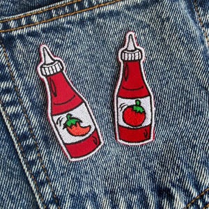 May include: Two embroidered patches in the shape of ketchup bottles, with red and white details. One features a chili pepper, the other a tomato. The patches are set against a blue denim background.