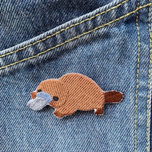 Puede incluir: Un parche bordado de un ornitorrinco marrón con un pico azul sobre un fondo de mezclilla azul.
