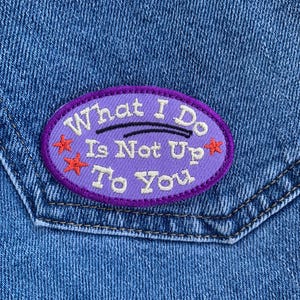 Puede incluir: Primer plano de un bolsillo de mezclilla con un parche bordado morado. El parche tiene el texto "What I Do Is Not Up To You" en letras blancas, con acentos de estrellas naranjas.