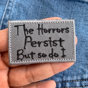 Puede incluir: Un parche rectangular gris con el texto "The Horrors Persist But so do I" en letras negras. El parche tiene un borde cosido y está sobre un fondo de mezclilla azul.