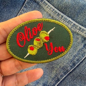 以下が含まれることがあります： 「Olive You」の赤い文字が刺繍された楕円形のパッチ。赤い中心を持つ3つの緑色のオリーブが串に刺されており、金色の縁取りのある濃い緑色の背景に配置されています。パッチはデニムの背景にあります。
