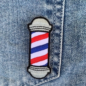 以下が含まれることがあります： 黒の輪郭のある赤、白、青の理髪店のポールパッチ。パッチは青いデニムの背景に縫い付けられています