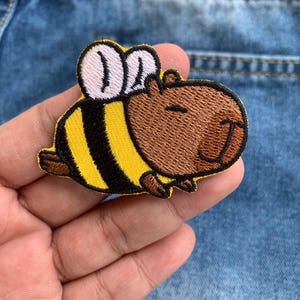 May include: Embroidered patch featuring a capybara with bee wings and a striped yellow and black body. The capybara has a brown embroidered face with a smiling expression. The patch is on a blue denim background.
