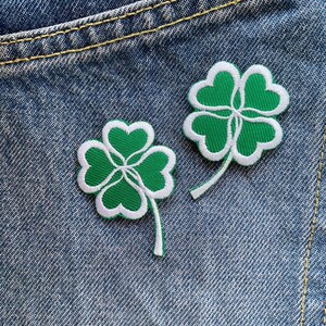 Könnte beinhalten: Zwei grüne vierblättrige Kleeblatt-Patches mit weißen Umrandungen, die auf einen blauen Jeansstoff genäht sind.