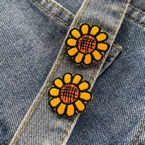 Könnte beinhalten: Nahaufnahme von Jeansstoff mit zwei gestickten Sonnenblumen-Patches. Die Sonnenblumen haben gelbe Blütenblätter, orangefarbene Zentren und schwarze Umrisse. Der Denim hat sichtbare Nähte und eine hellblaue Waschung.