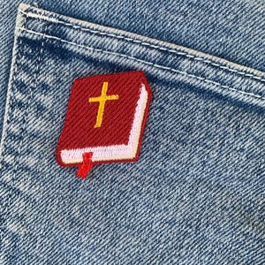 May include: A red Bible patch with a gold cross and a white border is sewn onto a blue denim pocket. The patch has a red bookmark. The denim has a light blue wash.