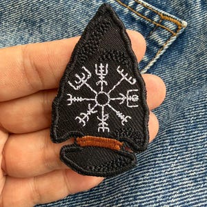 May include: Black arrowhead patch featuring a white rune symbol and a brown accent. The patch has an embroidered design. This patch is likely for clothing or accessories.