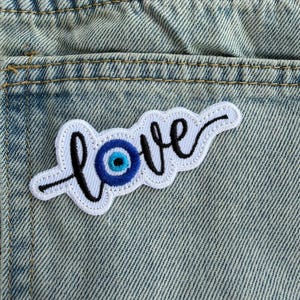 May include: An embroidered patch featuring the word love in black cursive script, with the letter o replaced by a circular blue, white, and black evil eye symbol. The patch has a white border and is attached to light blue denim fabric.