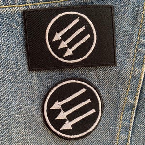Puede incluir: Dos parches de tela negra con flechas blancas bordadas en un círculo. El parche superior es rectangular y el parche inferior es redondo.