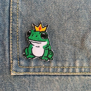 Może przedstawiać: Zielona żaba w złotej koronie, wyszyta na niebieskim denimie.