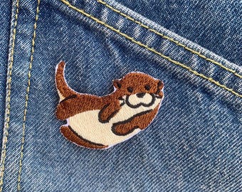 Può includere: Una toppa ricamata di una lontra che nuota, in colori marrone e crema, su uno sfondo di denim blu.