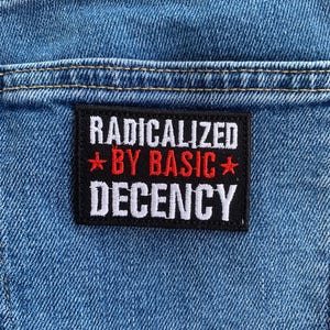May include: Close-up of a blue denim fabric with a black rectangular patch. The patch features the text "RADICALIZED" in white, "BY BASIC" in red, and "DECENCY" in white, with red star accents.