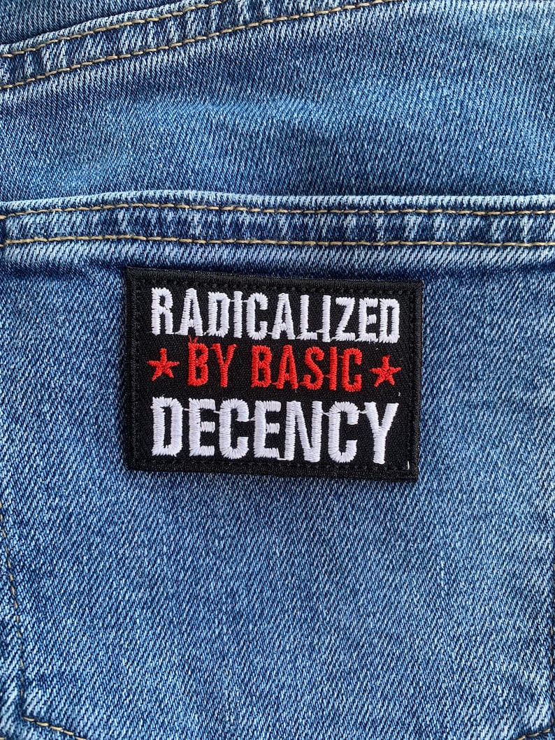 May include: Close-up of blue denim fabric with a black rectangular patch. The patch features the text "RADICALIZED" in white, "BY BASIC" in red, and "DECENCY" in white, with red star accents.
