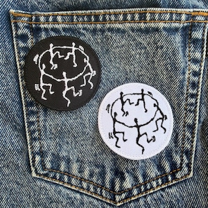 May include: Two round patches with a black and white design of stick figures in a circle are displayed on a denim pocket. One patch is black with white figures, and the other is white with black figures. The patches are embroidered.