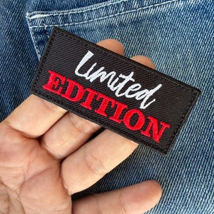 Peut inclure: Un écusson rectangulaire noir avec les mots "Limited Edition" brodés en blanc et rouge. L'écusson est posé sur un fond en denim bleu, mettant en valeur la texture du tissu.