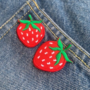以下が含まれることがあります： 青いデニムの背景に、緑の葉が付いた赤い刺繍のイチゴのパッチが2つある。