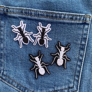 以下が含まれることがあります： 青いデニムのポケットに、白と黒の刺繍されたアリのパッチが4つ配置されています。パッチは詳細なデザインで、アリの体と脚がはっきりと定義されています。デニム生地にはステッチが見えます。
