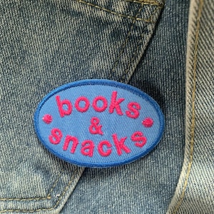 Op de afbeelding: Blauw en roze geborduurd patch met de tekst "books & snacks" op een denim achtergrond.