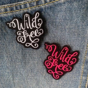 Peut inclure: Deux écussons brodés avec les mots "Wild Free" en écriture cursive. L'un a un texte blanc sur fond noir, l'autre un texte rose sur fond noir. Les écussons sont posés sur un fond en jean.