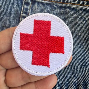 May include: A round, white embroidered patch featuring a bold red cross in the center. The patch is held against a denim background, showcasing the detailed stitching and vibrant colors. This patch is a symbol of aid and assistance.