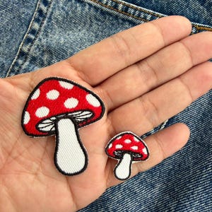 以下が含まれることがあります： 2つのキノコのパッチ。大きなパッチは、白い水玉模様の赤い帽子、白い茎、黒いアウトラインがあります。小さなパッチはデザインを反映しています。どちらも青いデニムの背景にあります。