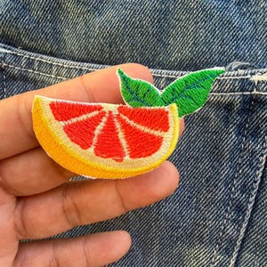May include: Embroidered patch featuring a grapefruit slice with red segments, a yellow rind, and two green leaves. The patch is designed to be ironed or sewn onto fabric, adding a decorative element to clothing or accessories.