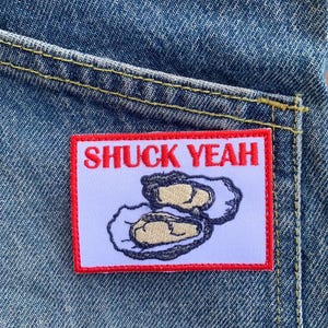 以下が含まれることがあります： デニムポケットのクローズアップ。長方形のパッチが付いており、赤い縁取りがあり、赤い文字で「SHUCK YEAH」と刺繍されたカキのデザインが描かれています。