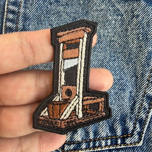Könnte beinhalten: Gestickter Aufnäher, der eine Guillotine in Braun-, Schwarz- und Weißtönen darstellt. Der Aufnäher hat einen schwarzen Rand und ist zum Aufbügeln oder Aufnähen auf Stoff vorgesehen. Das Design ist detailliert und zeigt die Komponenten der Struktur.