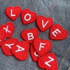 Może przedstawiać: Czerwone naszywki w kształcie serca z białymi haftowanymi literami, w tym słowo "LOVE". Naszywki są rozrzucone na niebieskim tle z dżinsu. Idealne do projektów DIY, takich jak dekorowanie odzieży lub akcesoriów.