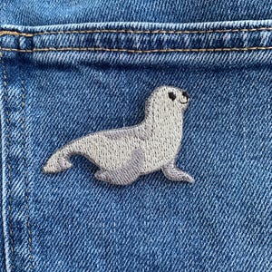 以下が含まれることがあります： 黒い目と笑顔の表情を持つ灰色のオットセイの刺繍パッチ。パッチは、ポケットの近くの青いデニム生地に縫い付けられています。オットセイは横たわっています。