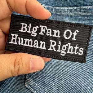Può includere: Una toppa rettangolare nera con testo ricamato bianco che recita "Big Fan Of Human Rights". La toppa è mostrata su uno sfondo di denim blu, evidenziando il messaggio e il design della toppa.