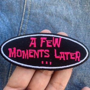Może przedstawiać: Czarna owalna naszywka z białym obramowaniem i różowym napisem "A FEW MOMENTS LATER". Naszywka jest prezentowana na tle dżinsu.