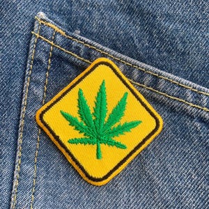 Peut inclure: Un écusson jaune en forme de losange avec une bordure noire et un motif de feuille de cannabis verte. L'écusson est cousu sur une poche en jean bleu, avec des coutures jaunes visibles.