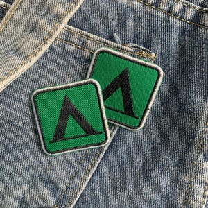 May include: Two green and white embroidered patches with a black tent symbol. The patches are on a blue denim background.