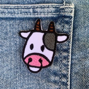 Op de afbeelding: Een witte koe-patch met zwarte vlekken en een roze neus, genaaid op een blauwe denim achtergrond.