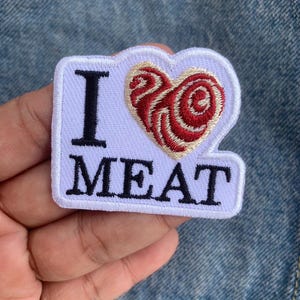 May include: White embroidered patch with the words "I MEAT" in black block letters. A red and tan heart design is above the word "MEAT". The patch is rectangular with a white border.
