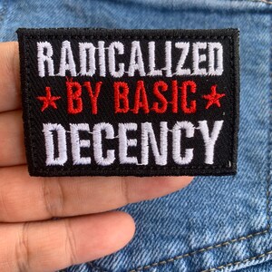 May include: A black rectangular patch with the words "RADICALIZED BY BASIC DECENCY" in white and red. The patch is held against a blue denim background.