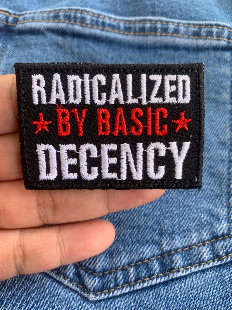 May include: A black rectangular patch with the words "RADICALIZED BY BASIC DECENCY" in white and red. The patch is held against a blue denim background.