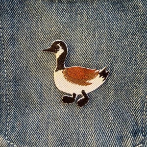 May include: Embroidered Canada goose patch with a black head and neck, white cheek, and brown body. The patch is attached to a blue denim pocket. The goose design is detailed and the patch is well-stitched.
