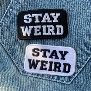 Op de afbeelding: Twee strijklabels met de tekst "Stay Weird" in witte letters op een zwarte achtergrond en zwarte letters op een witte achtergrond.