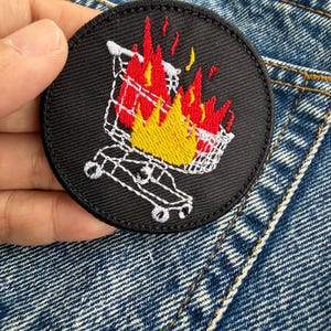 Puede incluir: Un parche redondo y negro bordado con un carrito de la compra blanco en llamas. Las llamas están representadas en tonos rojos y amarillos. El parche está diseñado para ser planchado o cosido en ropa o accesorios.